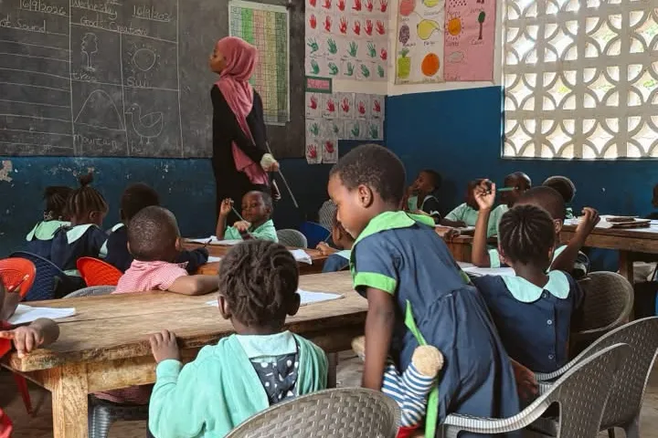 Alumnos en pupitres de madera siguiendo una clase mientras la maestra señala el pizarrón